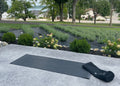 A black yoga mat and matching black rolled towel or mat cover placed on a light gray stone patio, with a landscaped garden, trees, and houses in the background.